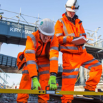 Two people dressed in orange high visibility PPE take a measurement on a work site